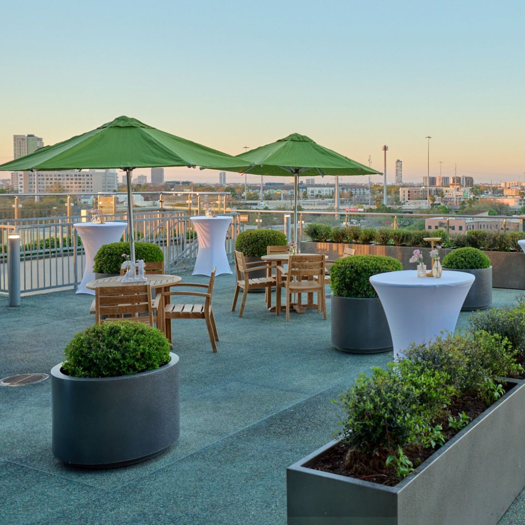 A modern outdoor rooftop terrace and event venue at Museo Houston, Panama City, set up for a private social gathering. The patio features wooden tables, cocktail tables with white linens, large lime green umbrellas, potted topiary plants, and a glass railing overlooking the city skyline at dusk