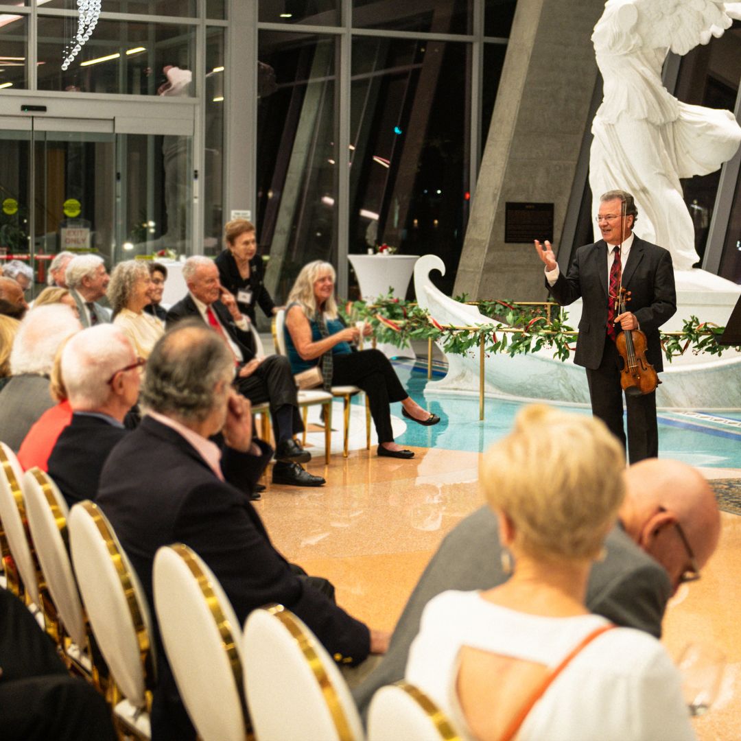A guest speaker presenting to a seated audience in front of the classical Kouros statue in the elegant Museo lobby.