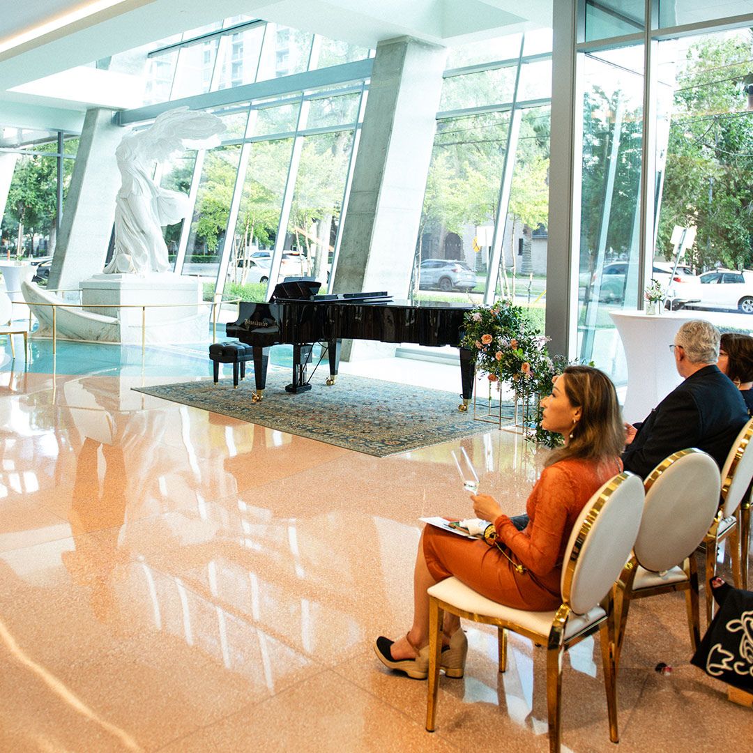 A grand piano positioned within the modern architectural setting of the Museo Houston entrance.
