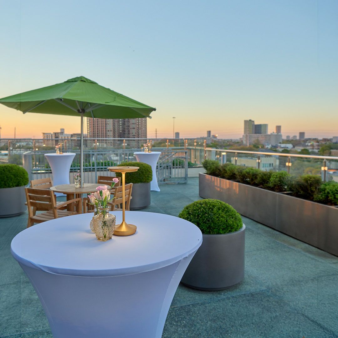 A white cocktail table with a green umbrella on the Museo Houston terrace, overlooking the city skyline at sunset.